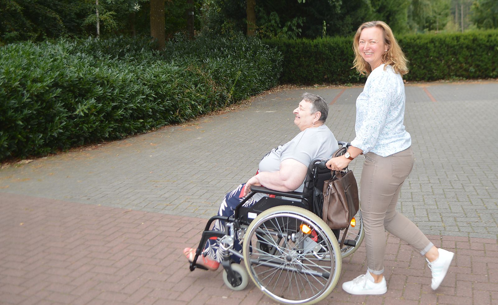 Svetlana Heidt schiebt eine lächelnde Frau im Rollstuhl über einen ruhigen, von Hecken gesäumten Platz